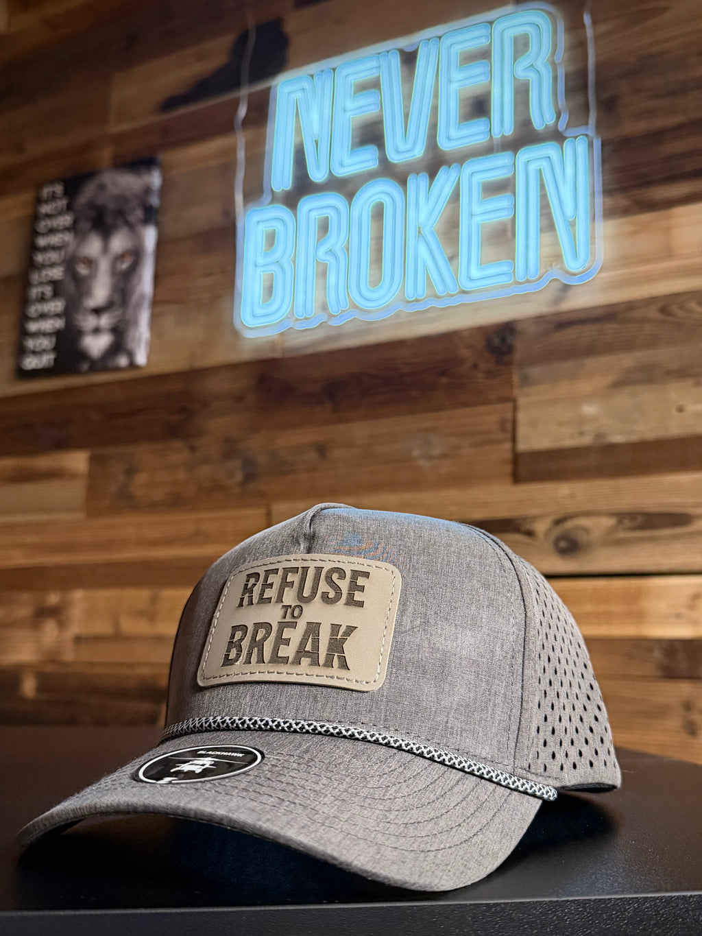 Gray cap with 'Refuse to Break' patch on a wooden background with 'Never Broken' neon sign.